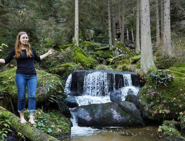 Biologin und Wildkraeuterguide Gerda Holzmann ist gerne in den Waeldern des Waldviertels unterwes und balanciert hier auf einem Baumstamm im Wald, neben einem Wasserfall.