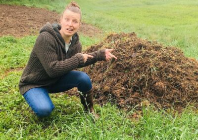 Kräuterfrau Gerda Holzmann kniet in der Wiese vor einem großen Haufen Queckenwurzel, die sie händisch aus einem Acker entfernt hat und zieht eine Grimasse.